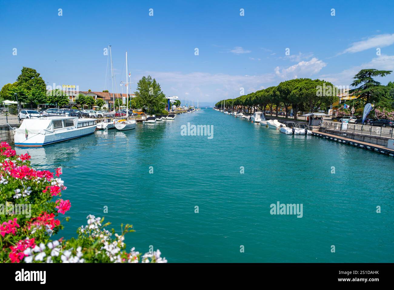 Malerischer Blick auf einen Kanal, der von Booten und Yachten gesäumt ist, umgeben von üppigem Grün, farbenfrohen Blumen und einer von Bäumen gesäumten Promenade unter einem klaren blauen Himmel. Stockfoto