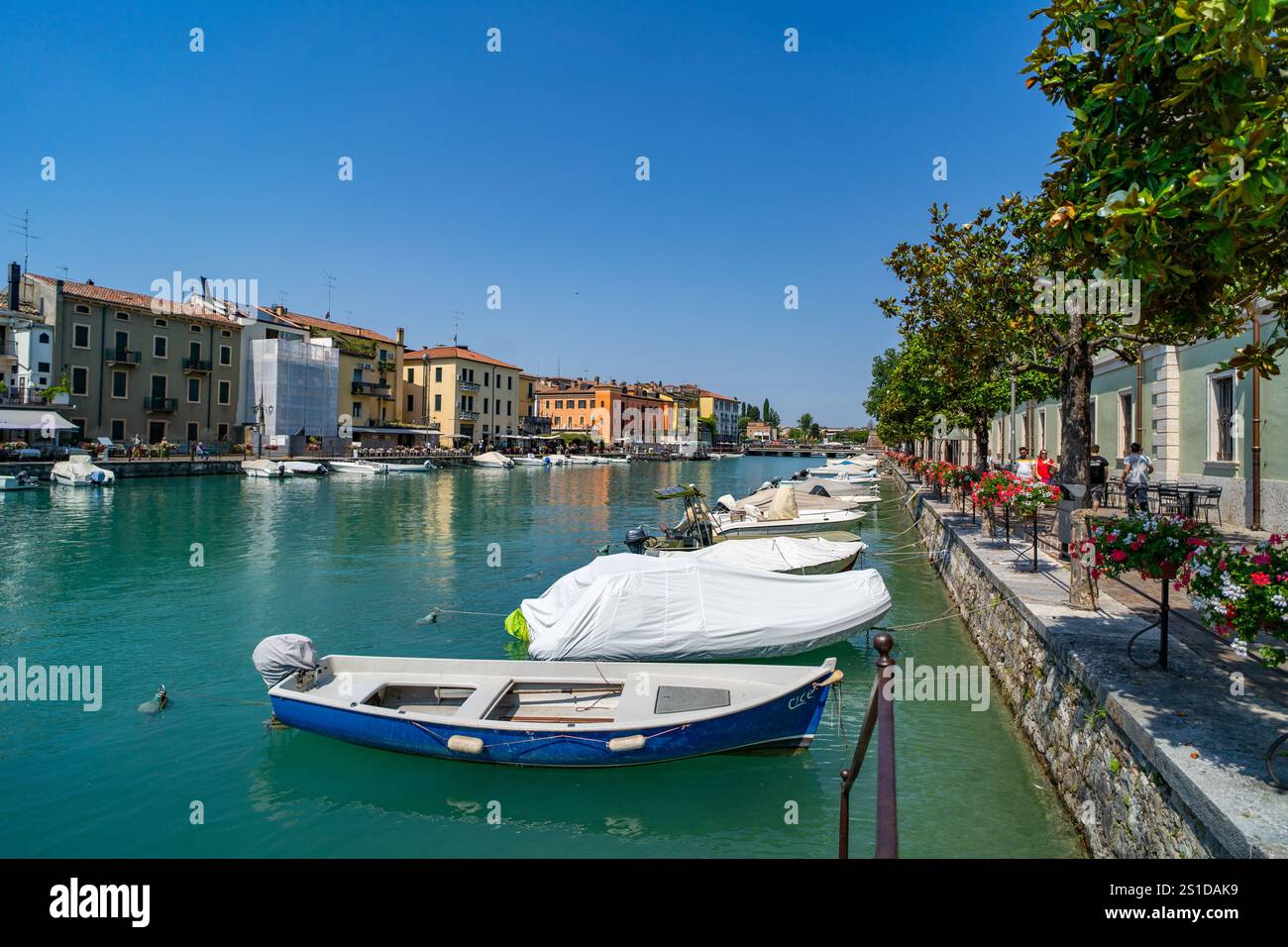 Malerische Kanalszene in einer europäischen Stadt mit farbenfrohen Gebäuden, vertäuten Booten und einer von Bäumen gesäumten Promenade mit leuchtenden Blumen. Ruhiges Wasser Stockfoto