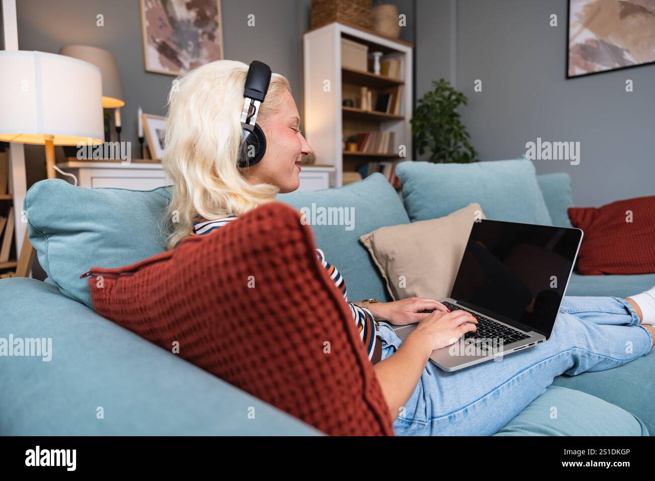 Junge lächelnde Frau mit schnurlosen Kopfhörern, die zu Hause ein Notebook benutzt. Freischaffende Hipster-Geschäftsfrau, die am Computer arbeitet, als Nebenbeschäftigung, passives Einkommen, Stockfoto