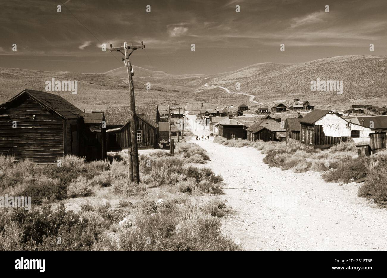 Staubige Hauptstraße von Bodie mit schiefen Holzhäusern, Lichtmasten und einem weiten Blick auf die umliegenden Präriehügel. Foto in Sepia-Ton. Staubige Hauptleitung Stockfoto