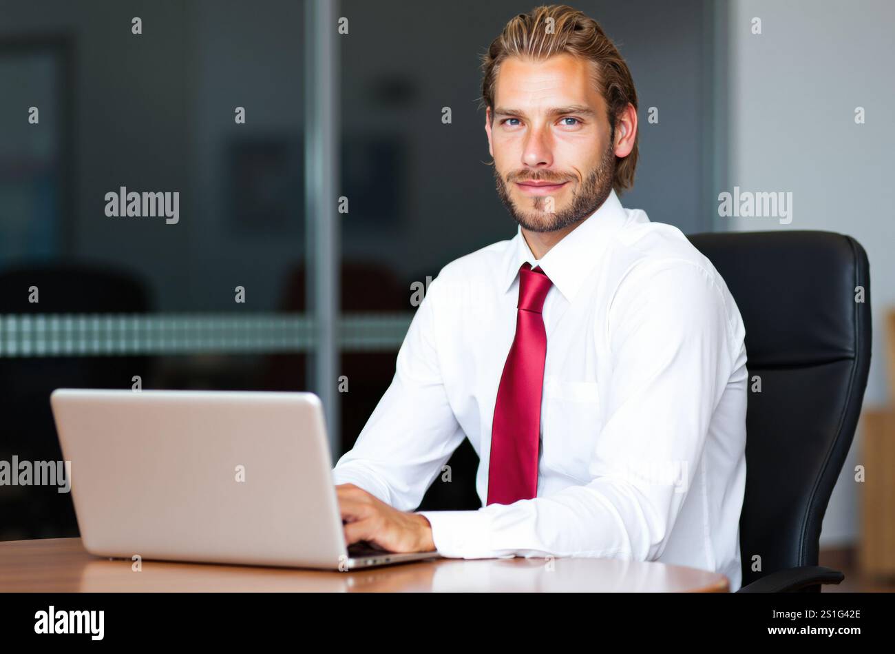 Junger Geschäftsmann, der an einem eleganten Schreibtisch sitzt und intensiv an seinem Laptop arbeitet, in einer modernen Büroumgebung, umgeben von einer hellen Inneneinrichtung und einem großen gl Stockfoto