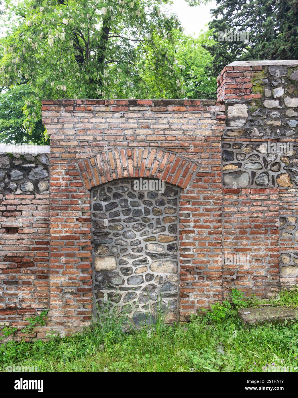 Eine verwitterte Mauer kombiniert Stein- und Ziegelmaterialien und verfügt über eine bogenförmige Nische. Üppiges Grün umgibt das Gebäude und unterstreicht seinen historischen Charme Stockfoto