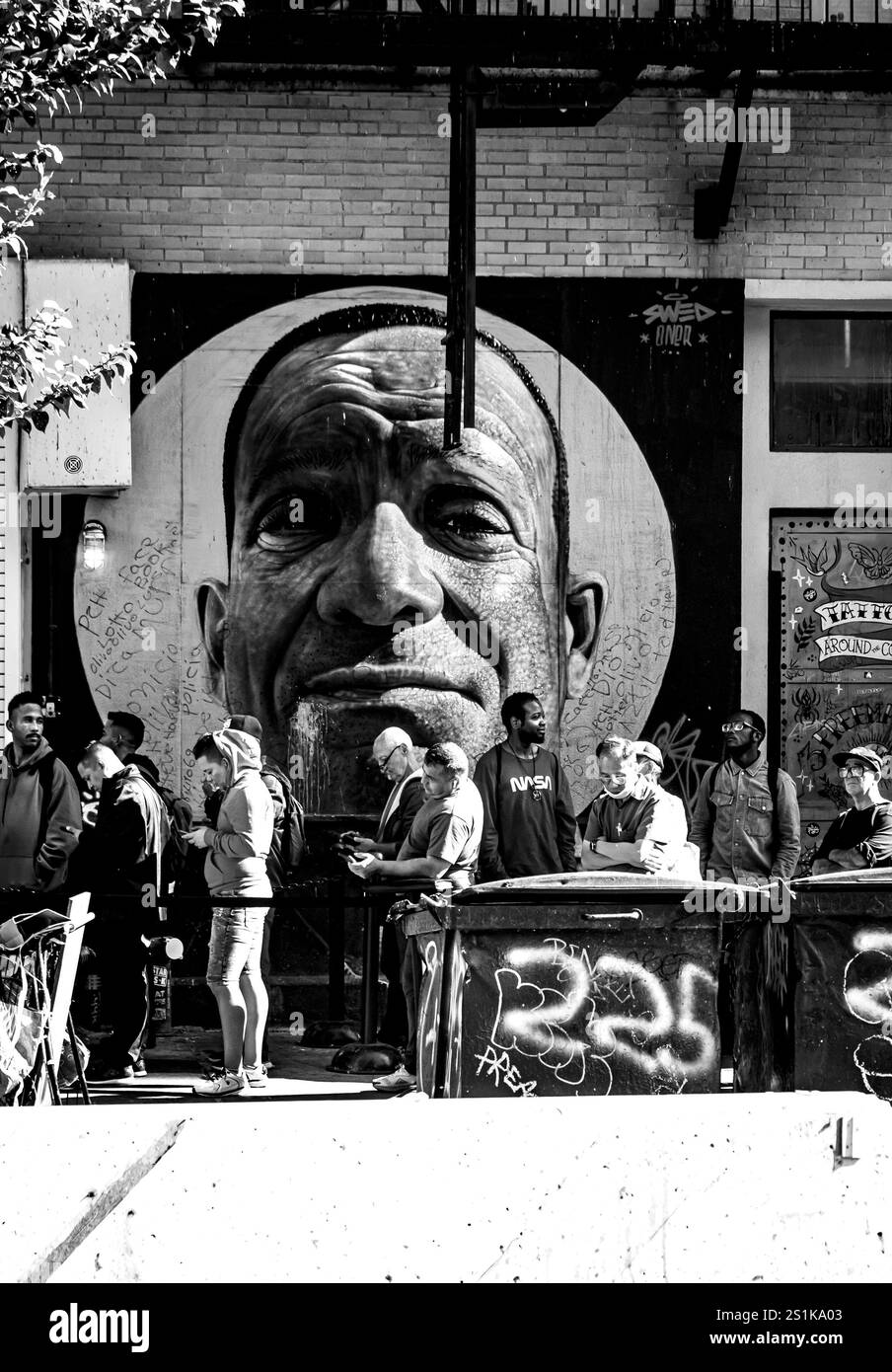 Eine Gruppe von Menschen steht in einer Reihe vor einem großen städtischen Wandgemälde, das das Gesicht eines Mannes im Street Art-Stil in Schwarz-weiß zeigt Stockfoto