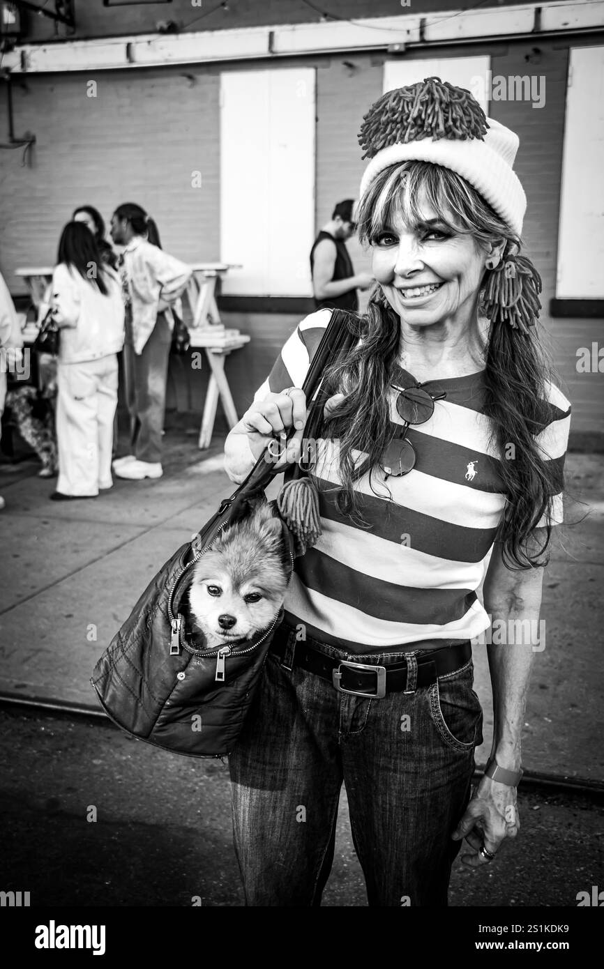 Lächelnde Frau in gestreiftem Hemd und Hut mit einem kleinen Hund in einer Tasche, stehend in einer lebhaften städtischen Umgebung. New York City Hundeparade, NYC, USA Stockfoto