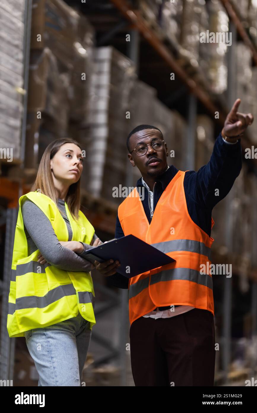 Ein Lagerverwaltungsteam, das sich auf betriebliche Aufgaben konzentriert. Zwei Arbeiter mit gut sichtbaren Westen stehen vor gestapelten Regalen Stockfoto