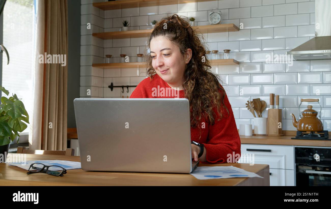 Eine fröhliche Frau in einer modernen Küche nutzt ihren Laptop und genießt einen entspannten Moment, während sie von zu Hause aus arbeitet oder studiert. Stockfoto