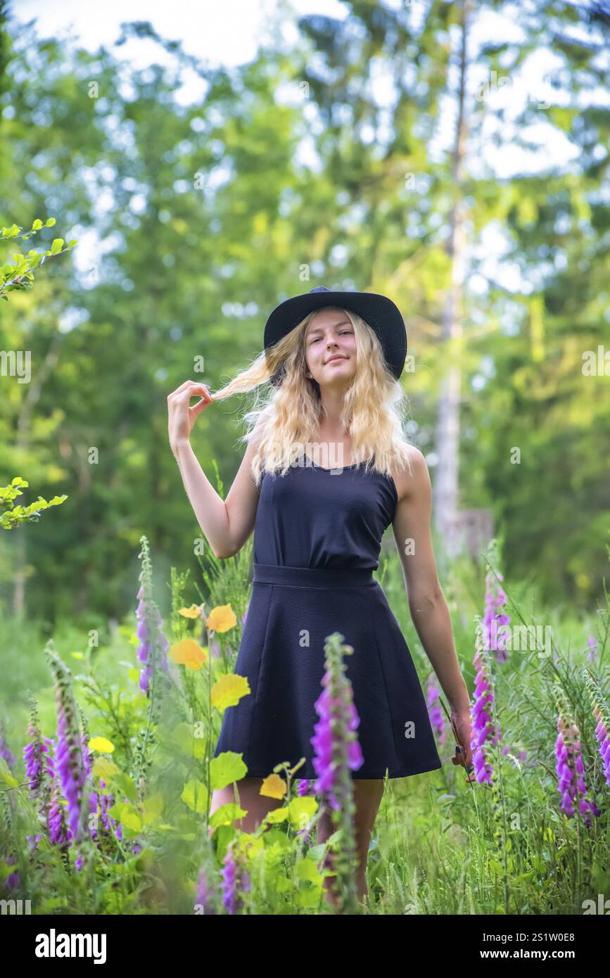 Junge blonde Frau mit schwarzem Kleid und Hut im Sommer mit Fuchshandschuhblumen im Waldportrait in der Natur. Junge blonde Frau mit schwarzem Kleid in Natur Stockfoto