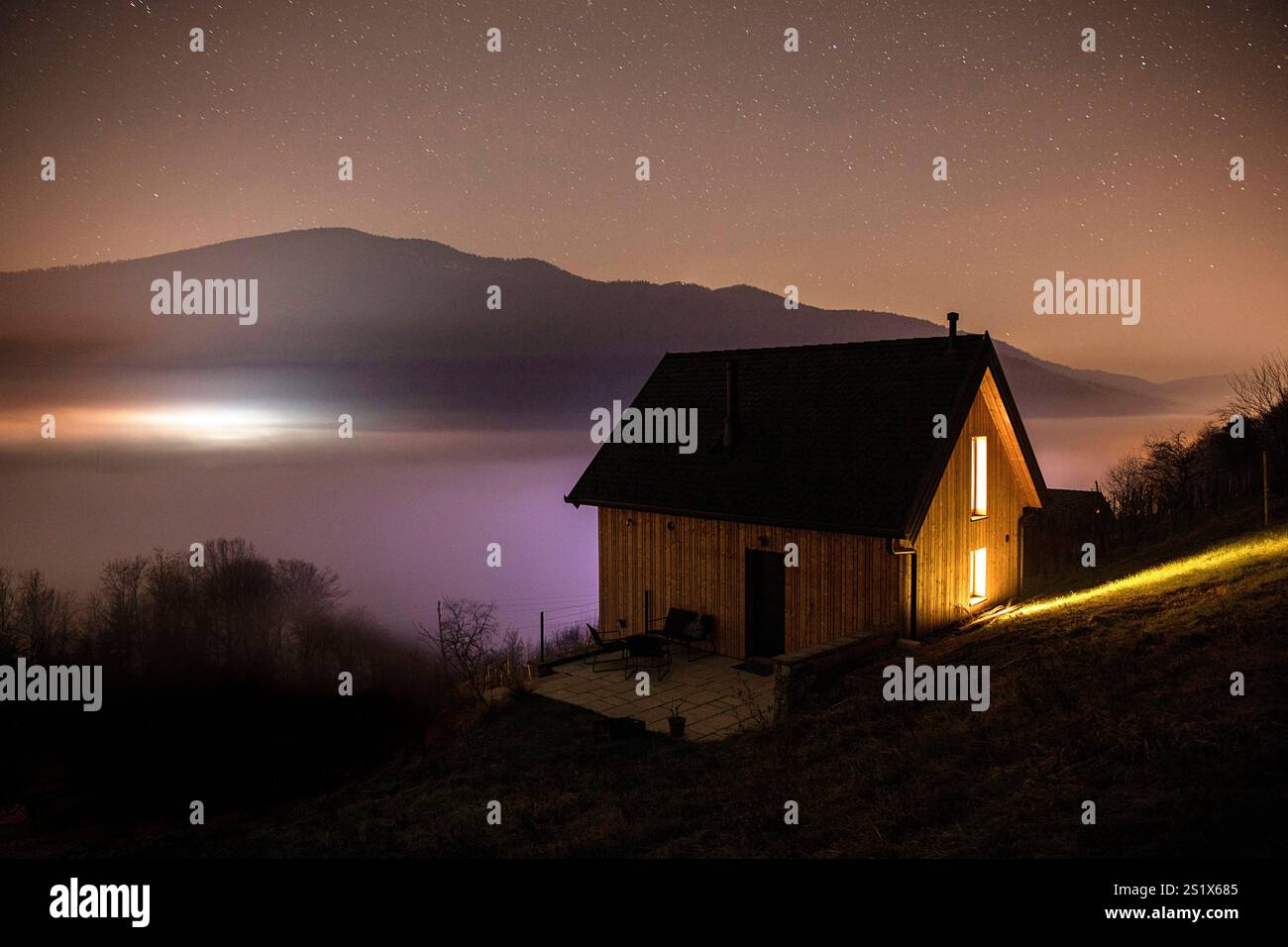 Schöne Holzhütte mit Lichtern in der Sternennacht, auf einem Hügel über dem Nebelmeer in der Weinlandschaft Dolenjske toplice, Slowenien Stockfoto