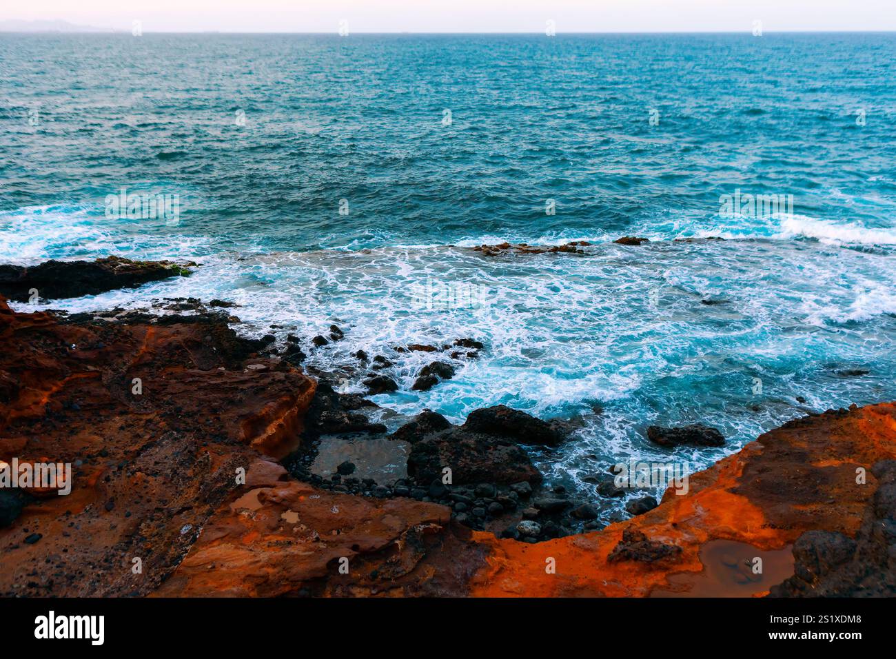 Felsige Küste mit Wellen, die gegen die Küste krachen. Wasser scheint eine Mischung aus Blau und weiß zu sein, da durch Wellen geschäumter Schaum entsteht. Zerklüftete Felsen Stockfoto