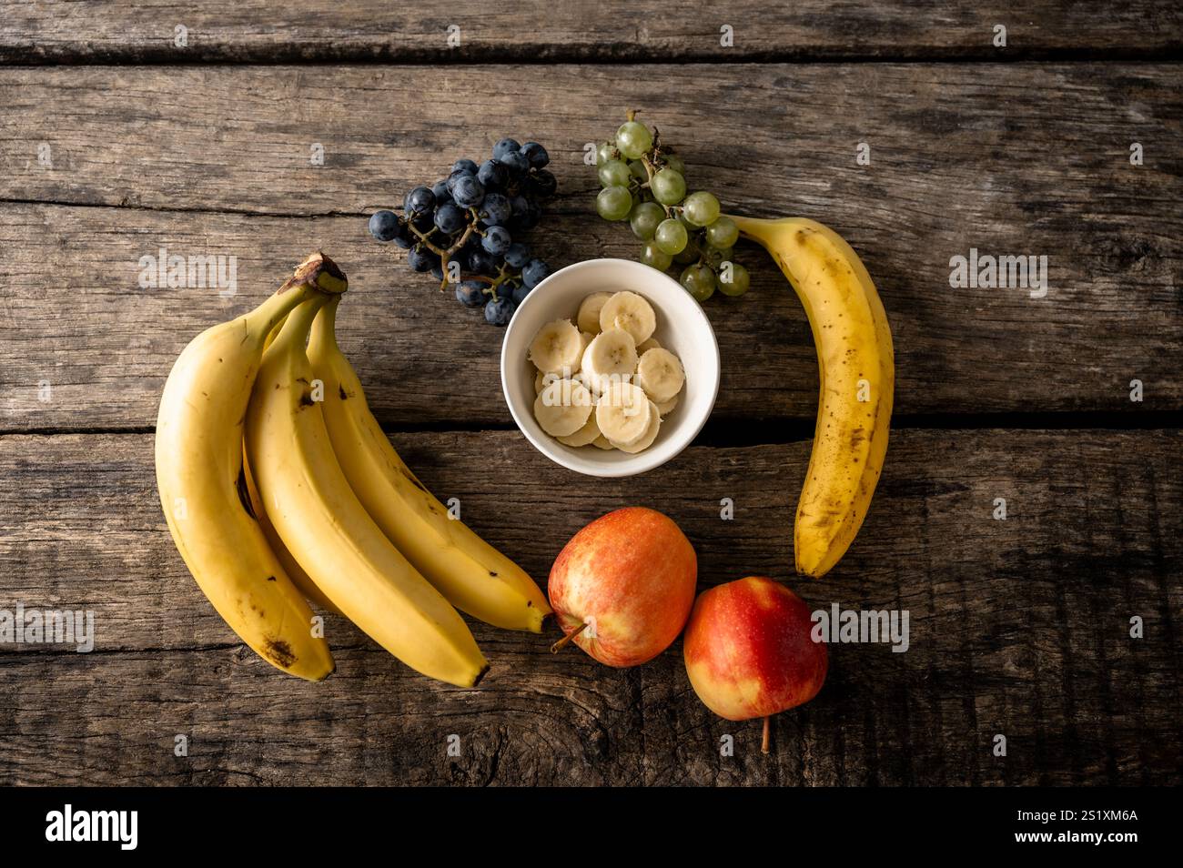 Eine Auswahl an Früchten, darunter Bananen, Äpfel, grüne und schwarze Trauben und eine Schüssel mit geschnittenen Bananen, die auf einer rustikalen Holzoberfläche angeordnet sind. Stockfoto