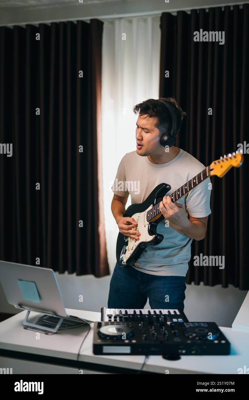 Asiatischer männlicher Produzent, der Gesang über elektronische Beats, Gitarre und Audio-Ausrüstung auf dem Schreibtisch in einer modernen und inspirierenden Studioumgebung überlagert. Stockfoto