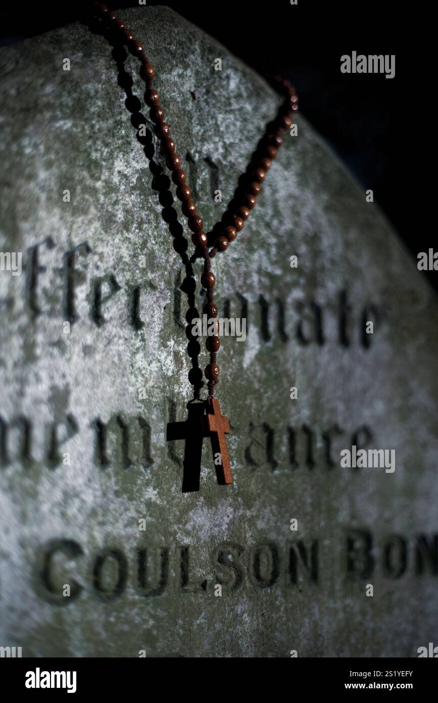 Alter verwitterter Grabstein mit Rosenkranz, in dunklem Licht getaucht. Vorstellung von Glauben, Zeit, gotischer Religion und unheimlicher Friedhofslandschaft irgendwo in England Stockfoto