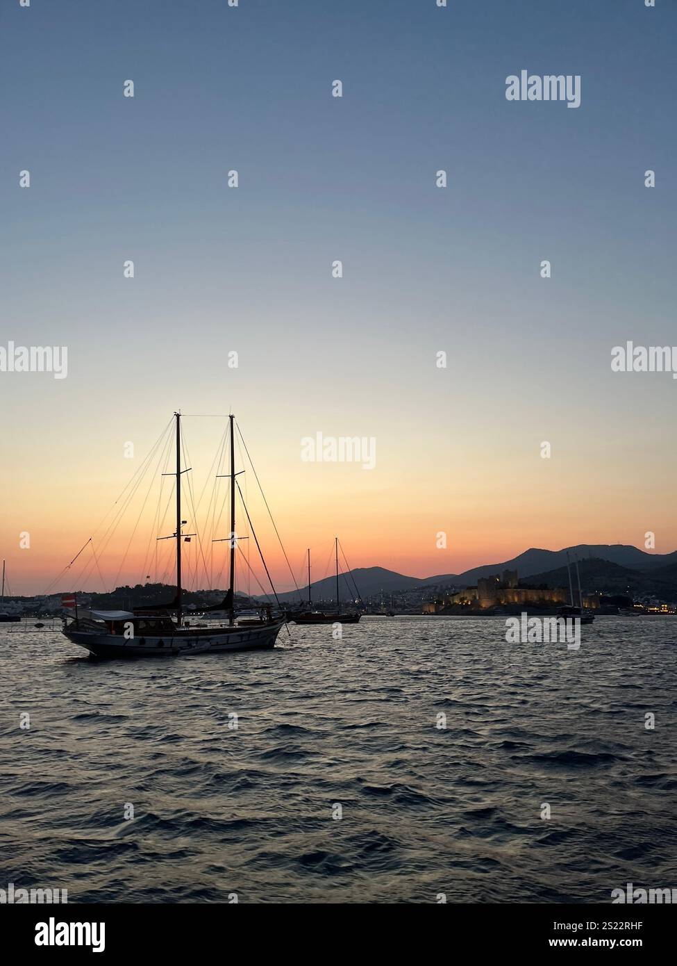 Eine wunderschöne vertikale Aufnahme der Yacht bei orangefarbenem Sonnenuntergang auf dem Meer mit Bergen im Hintergrund. Fantastische Yachtfahrt in Bodrum, Türkei. Stockfoto