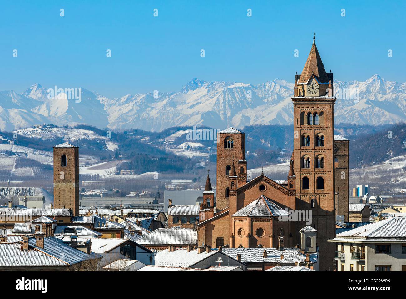 Stadt Alba mit schneebedeckten Dächern, mittelalterlichen Türmen und dem Kirchturm der Kathedrale vor schneebedeckten Alpen unter klarem blauem Himmel in Italien. Stockfoto