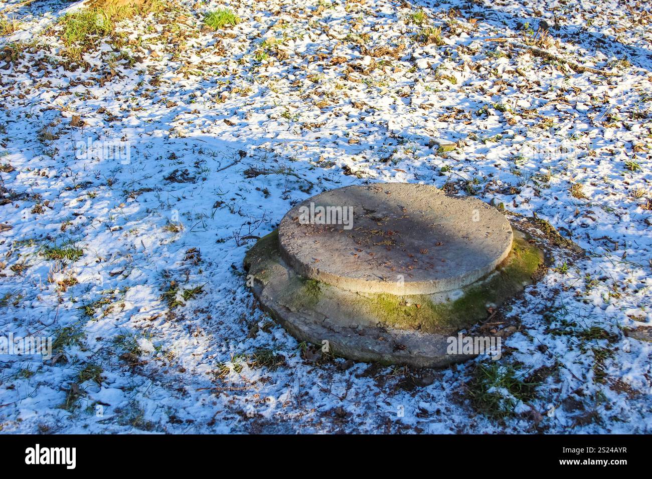 Ein runder Betonsockel liegt auf dem Boden auf, teilweise bedeckt von einer dünnen Schneeschicht an einem ruhigen Ort im Freien während des Winters. Stockfoto
