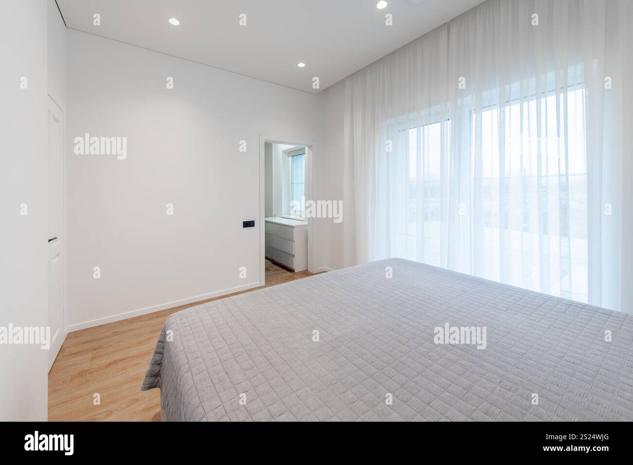 Das ruhige Schlafzimmer verfügt über Tageslicht durch große Fenster, minimalistische Einrichtung mit hellen Vorhängen und eine saubere Atmosphäre. Perfekt zum Entspannen Stockfoto
