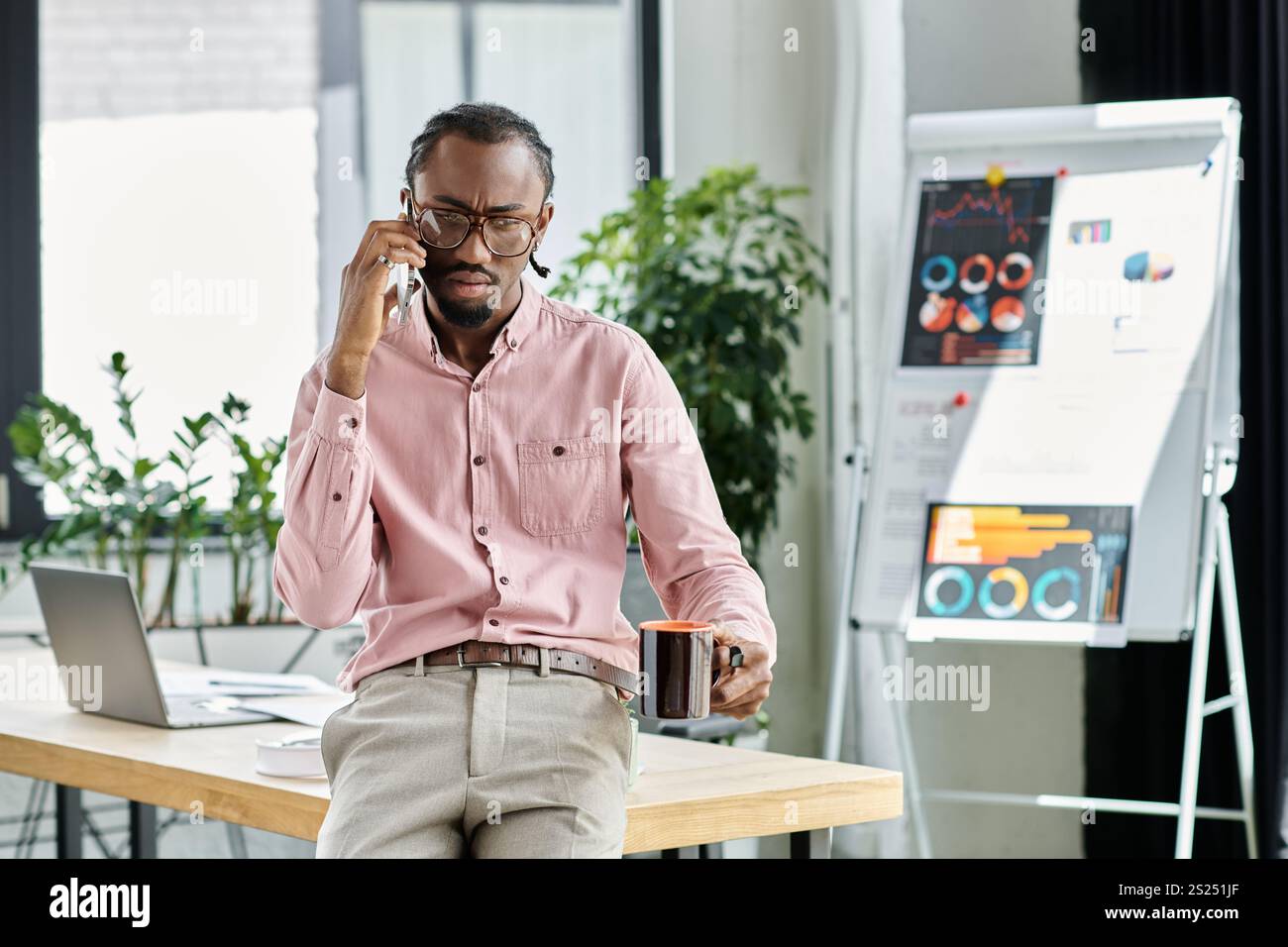 Ein fokussierter junger afroamerikanischer Mann, der während eines Anrufs mehrere Aufgaben erledigt, während er an einem schicken Arbeitsplatz Kaffee schlürft. Stockfoto