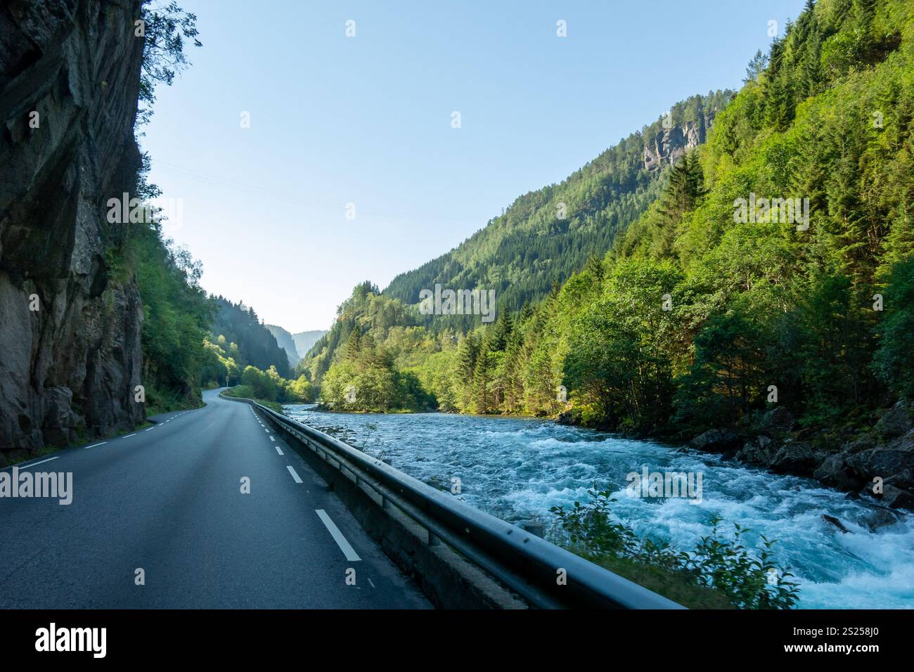 Eine gewundene Straße in Norwegen verläuft entlang eines lebendigen blauen Flusses, flankiert von hohen grünen Bäumen und steilen Bergen unter klarem Himmel Stockfoto