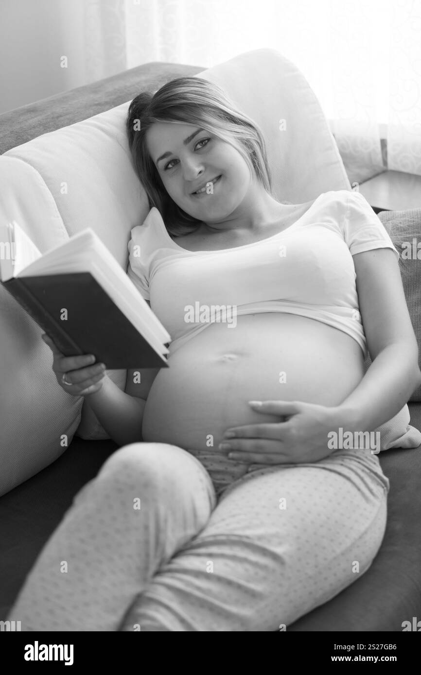 Schwarz-weiß-Bild einer schönen schwangeren Frau, die auf Sofa liegt und Buch liest Stockfoto