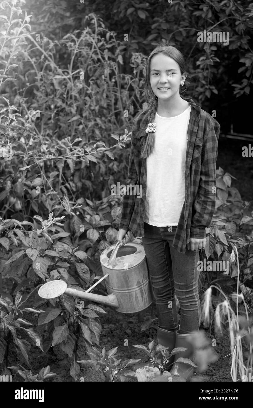 Schwarz-weiß-Bild eines glücklichen jungen Mädchens in roten Gummistiefeln, das Gemüse im Garten tränkt Stockfoto