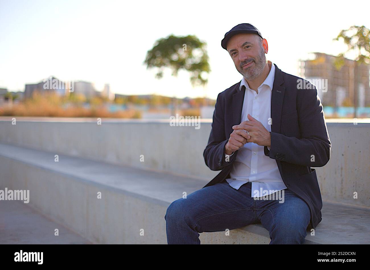 Mann, der draußen auf einer Betonbank sitzt, in einer urbanen Umgebung, in lässiger, aber professioneller Kleidung, die Selbstvertrauen ausstrahlt. Stadt im Hintergrund Stockfoto