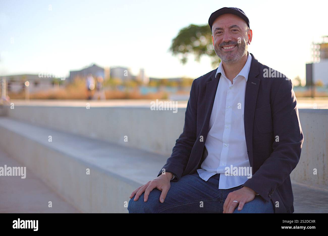 Ein fröhlicher Mann mittleren Alters in lässiger Kleidung sitzt auf einer Betonbank in einer hellen urbanen Umgebung im Freien, mit einer verschwommenen Stadtlandschaft im Hintergrund Stockfoto