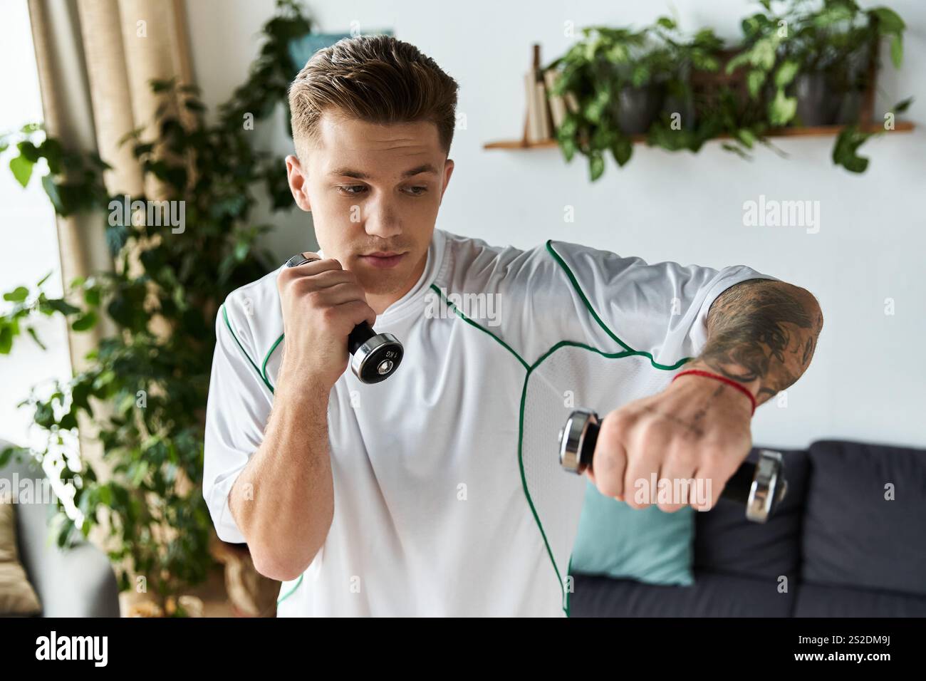 Der junge Mann trainiert drinnen und hebt Kurzhanteln mit einem festen Fokus auf Fitness. Stockfoto