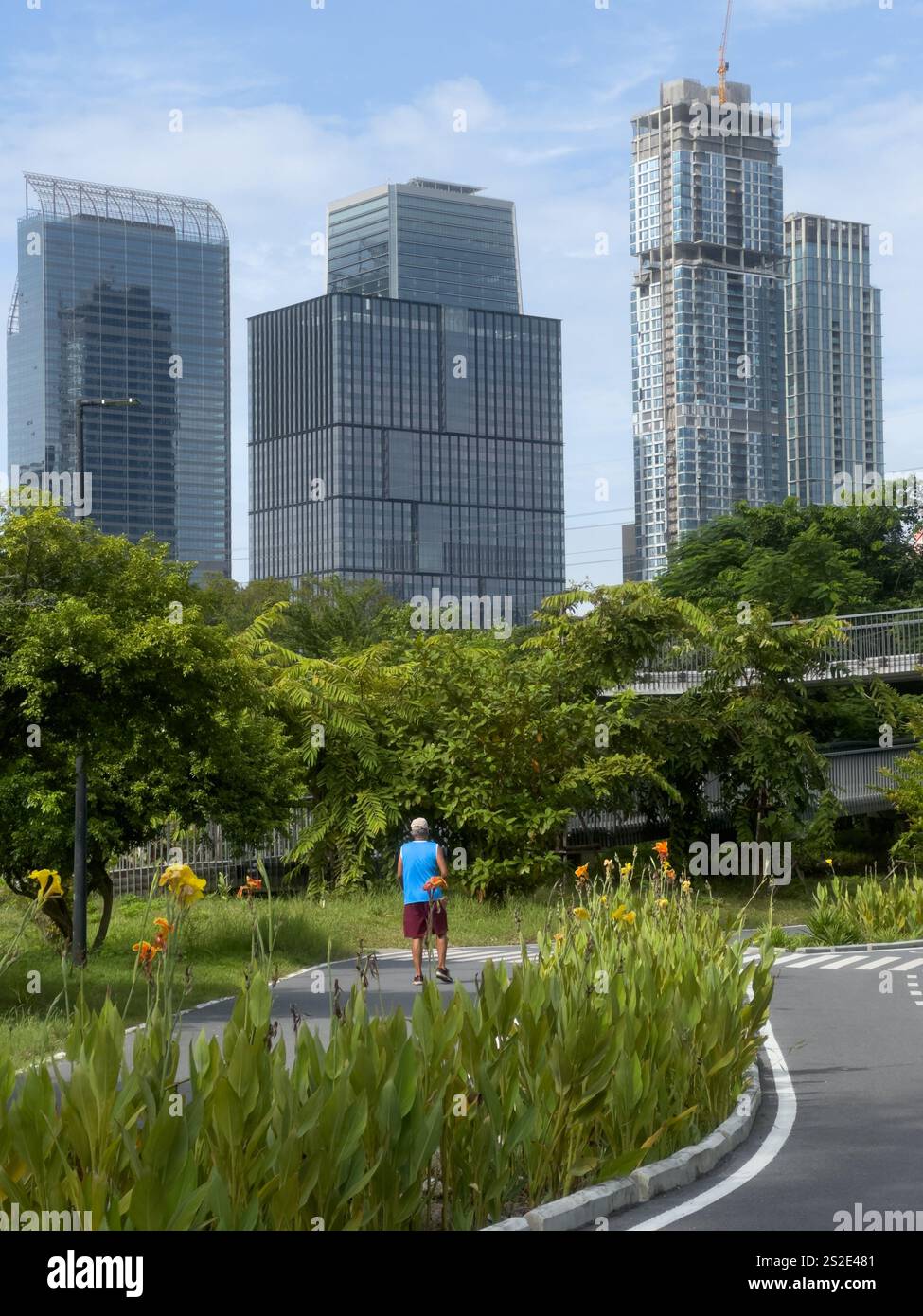Dieses Bild zeigt eine Person, die in einem üppigen Grün joggt, umgeben von hoch aufragenden modernen Wolkenkratzern in Bangkok, Thailand. Der ruhige Pfad gesäumt von Stockfoto