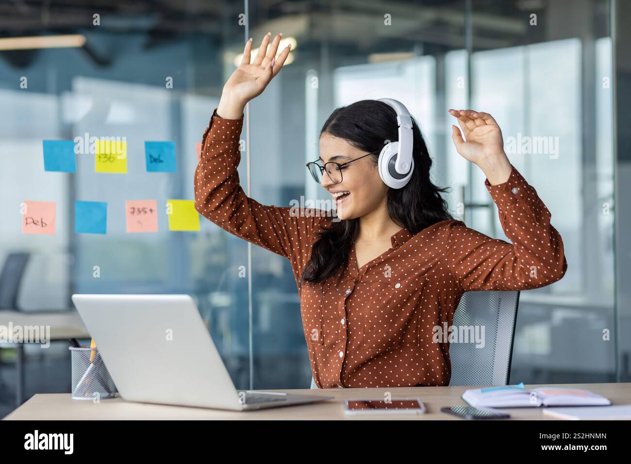 Fröhliche Frau, die Musik am Arbeitsplatz im Büro hört. Geschäftsfrau mit Kopfhörern, die sich am Laptop entspannen, tanzen und singen, während sie am Tisch sitzt. Stockfoto
