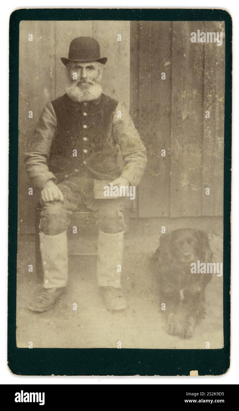 Original viktorianischer Carte de Visite (Visitenkarte oder CDV) viktorianischer Mann viktorianischer Gentleman. Der ältere Mann ist bärtig und aus dieser Kleidung ist ein Mann der Arbeiterklasse mittleren Alters, der einen Bowler-Hut und Lederleggings trägt, vor einem Bauernhaus sitzt, ein Buch hält, einen Arbeitshund an seiner Seite. Vielleicht ist der Mann ein Bauer oder Landarbeiter. War dieser Mann alphabetisch oder Analphabet - das Buch ist wahrscheinlich eine fotografische Requisite. Das Foto stammt von den 1870er bis Anfang der 1880er Jahre Stockfoto