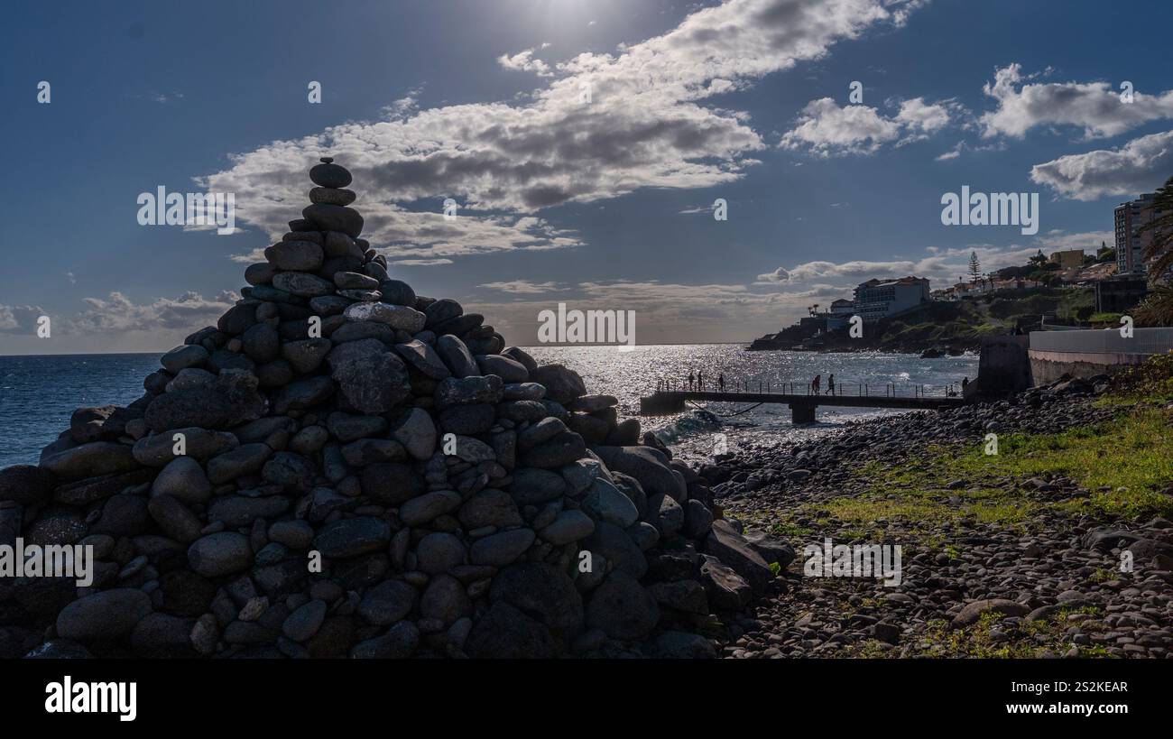 Eine Pyramide aus gestapelten Steinen an einem Kiesstrand bei Sonnenuntergang, mit einem Pier und entfernten Gebäuden, die sich vor dem Meer und dem bewölkten Himmel abheben. Stockfoto