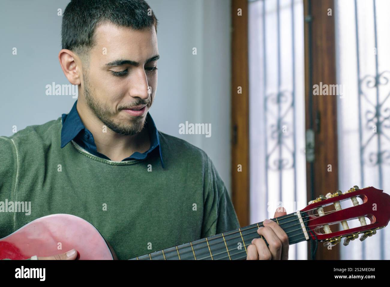 Junger Mann, der drinnen sitzt und eine rote Akustikgitarre spielt. Stockfoto Junger Mann, der drinnen sitzt und eine rote Akustikgitarre spielt. Stockfoto