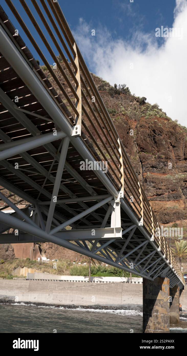 Eine moderne Fußgängerbrücke mit Metall- und Holzkonstruktion, die sich über eine felsige Küste mit einer Klippe vor einem klaren blauen Himmel erstreckt. Stockfoto