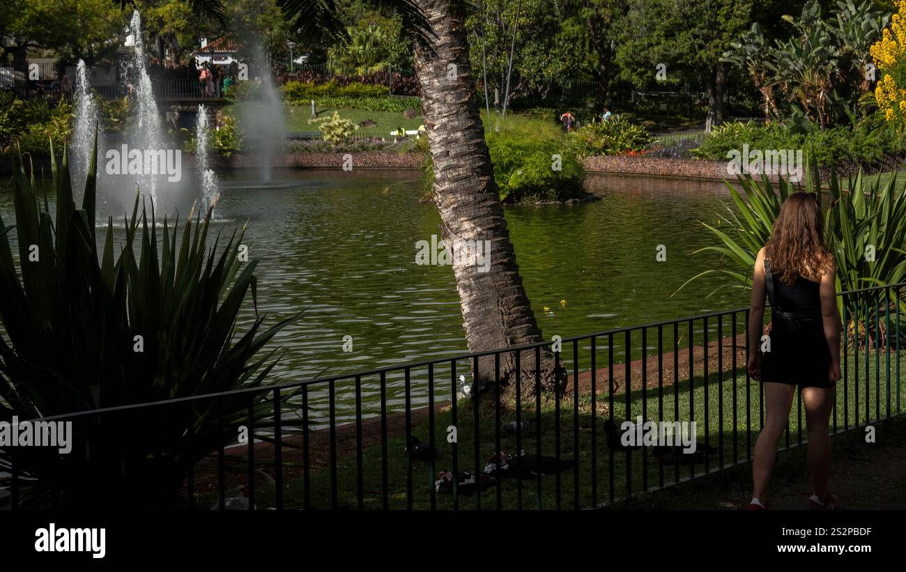 Ein landschaftlich reizvoller Park mit einem ruhigen Teich mit Springbrunnen, üppigem Grün und einer Frau, die an einem Geländer entlang läuft und die friedliche Umgebung im Freien genießt. Stockfoto