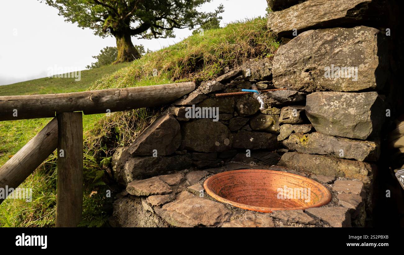 Ein rustikaler Wasserhahn im Freien mit einem Kupferbecken, in eine Steinmauer gebaut, in einer friedlichen ländlichen Landschaft mit einem Baum auf einem grasbewachsenen Hügel. Stockfoto