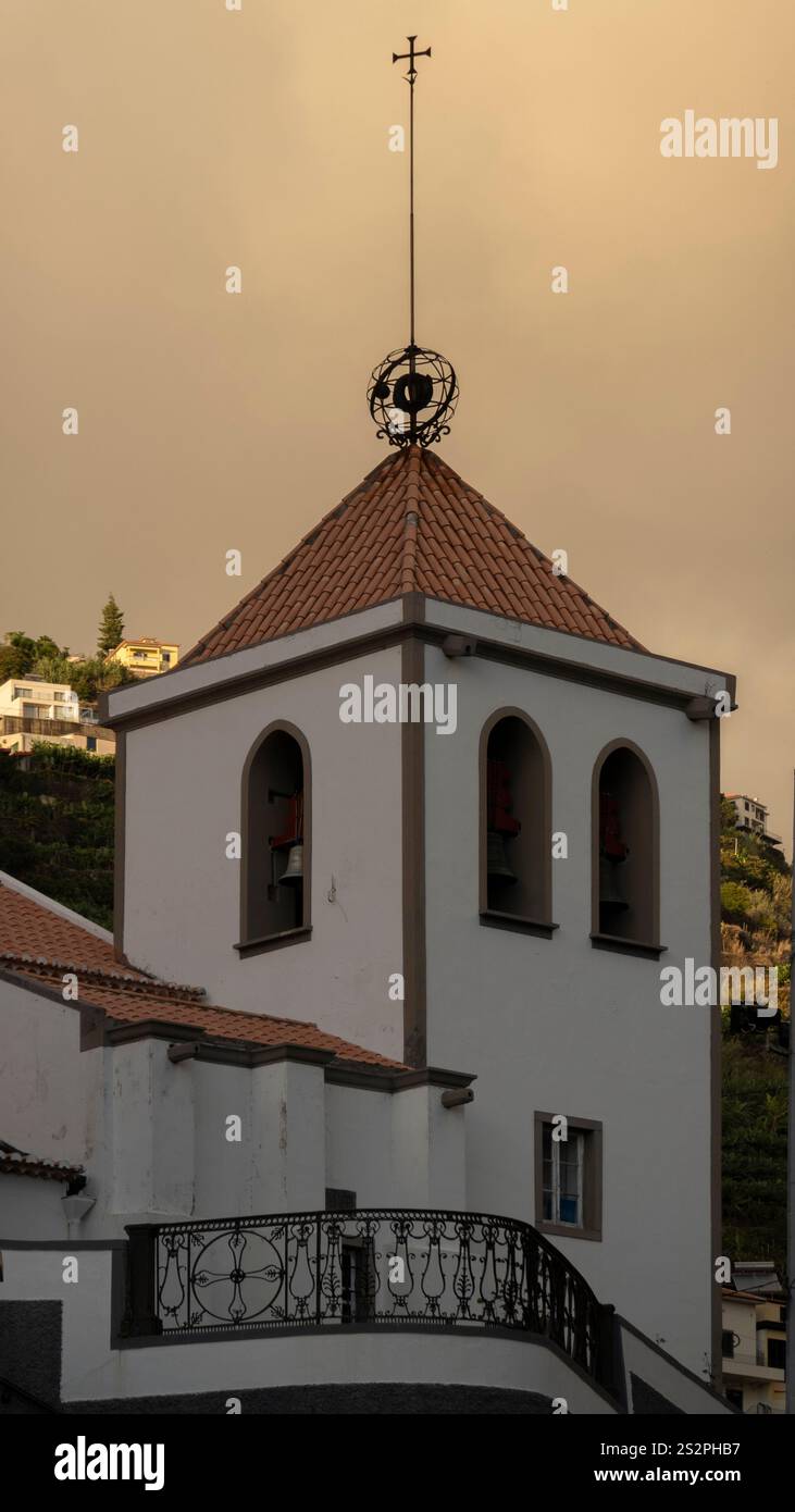 Ein Kirchturm mit Bogenfenstern und einem rot gekachelten Dach, das mit einem Kreuz geschmückt ist, vor einem goldenen Sonnenuntergangshimmel. Eine ruhige Architekturszene auf einem Hügel Stockfoto