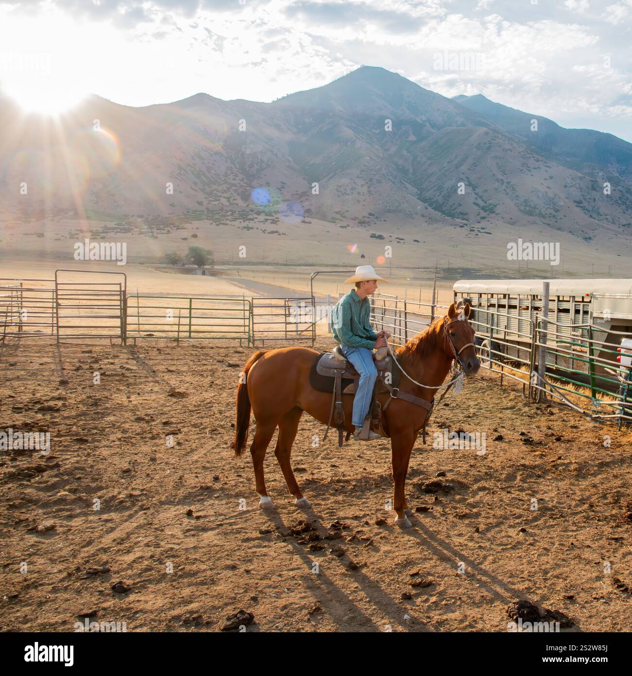 Ein Cowboy auf dem Pferd beobachtet einen Korral bei Sonnenaufgang, umgeben von Fechten und offenen Ebenen mit Bergen im Hintergrund. Die Sonne am frühen Morgen geht Stockfoto