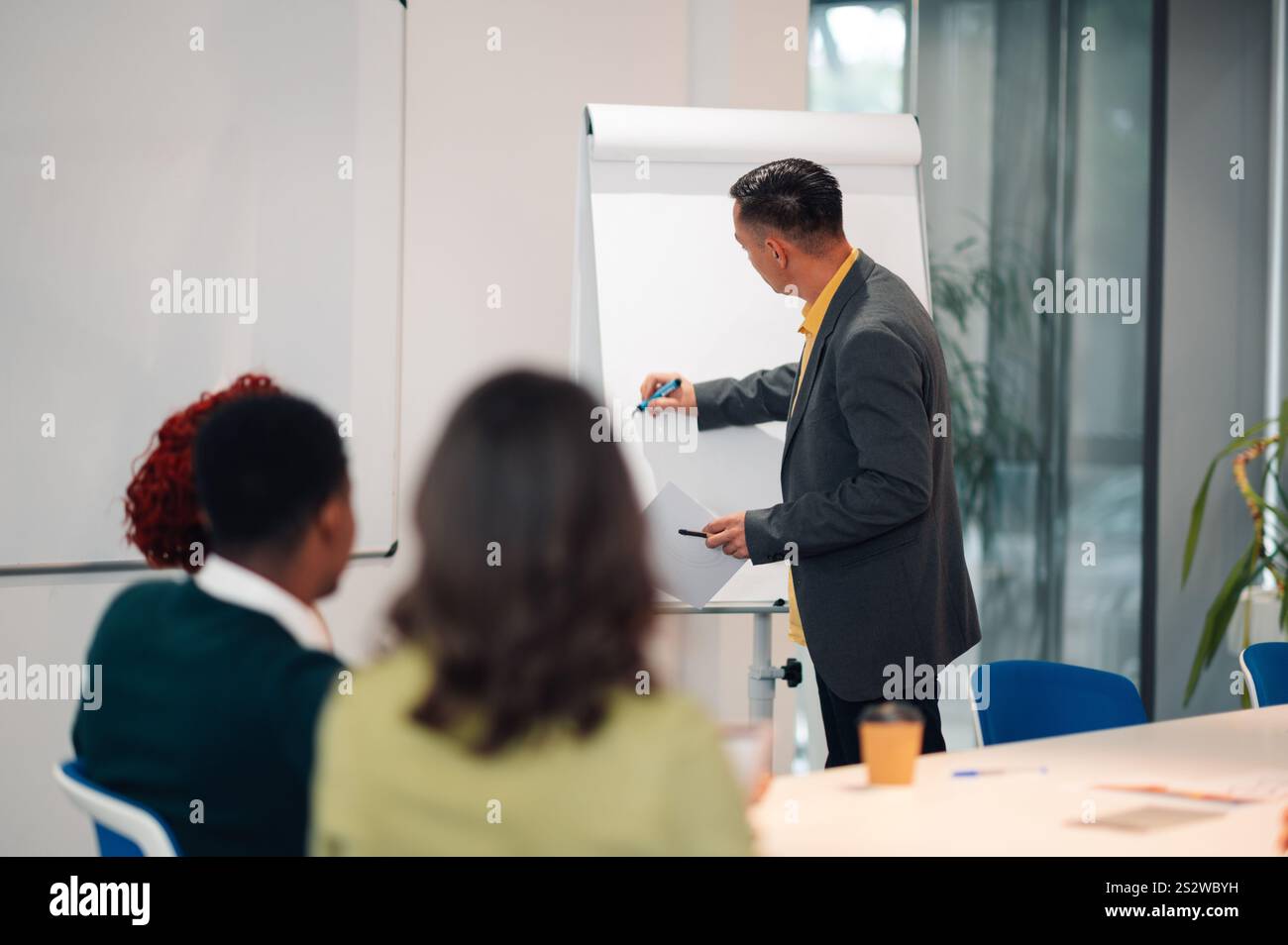 Rückansicht eines Geschäftsmannes mittleren Alters, der während einer Besprechung in einem Büro auf einem Whiteboard schreibt. Reifer männlicher Teamleiter zeichnet eine grafische Darstellung Stockfoto