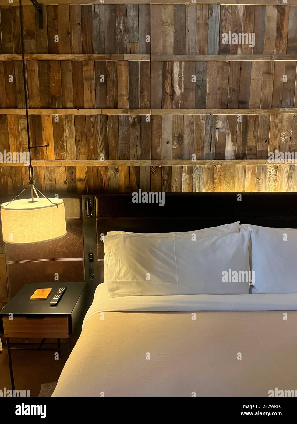 Ein gemütliches und stilvolles Schlafzimmer mit rustikaler Holzwand, einem ordentlich gefertigten Bett mit reinweißer Bettwäsche, das einen warmen, einladenden Glanz ausstrahlt. Minimalistisch - Smartphone-aufgenommenes Stockfoto