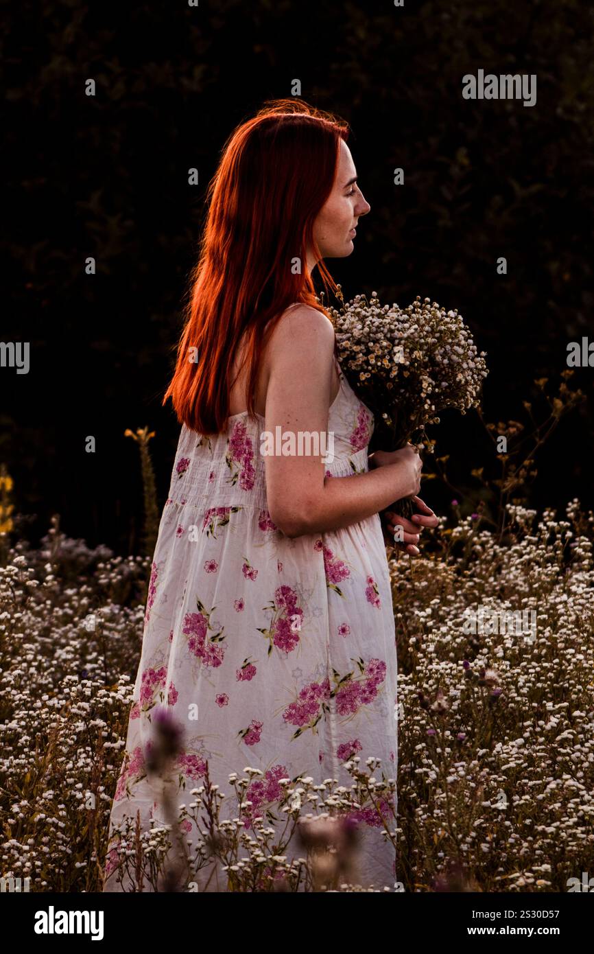 Ein rothaariges Mädchen steht in einem Gänseblümchenfeld, hält einen Blumenstrauß und blickt nachdenklich zur Seite Stockfoto