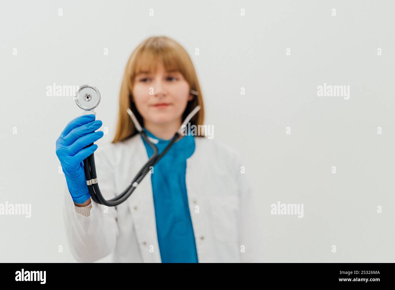 Eine Ärztin in einem weißen Mantel und blauen Handschuhen, die ein Stethoskop in Richtung Kamera hält. Medizinisches Fachpersonal im klinischen Umfeld Stockfoto