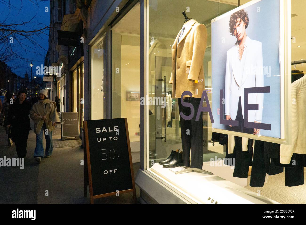 London, UK, 8. Januar 2025: Wenn die Dämmerung auf der Marylebone High Street einbricht, passieren die Käufer ein Schaufenster, in dem Herrenbekleidung ausgestellt wird und auf einer Tafel mit bis zu 50 % Rabatt auf den Januar-Verkauf geworben wird. Anna Watson/Alamy Live News Stockfoto London, UK, 8. Januar 2025: Wenn die Dämmerung auf der Marylebone High Street einbricht, passieren die Käufer ein Schaufenster, in dem Herrenbekleidung ausgestellt wird und auf einer Tafel mit bis zu 50 % Rabatt auf den Januar-Verkauf geworben wird. Anna Watson/Alamy Live News Stockfoto