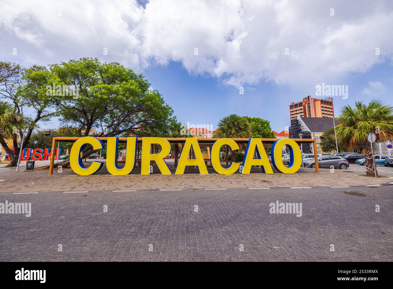 Großes gelbes Curacao-Schild im Freien mit Bäumen, Gebäuden und geparkten Autos im Hintergrund. Willemstad. Curacao. Stockfoto
