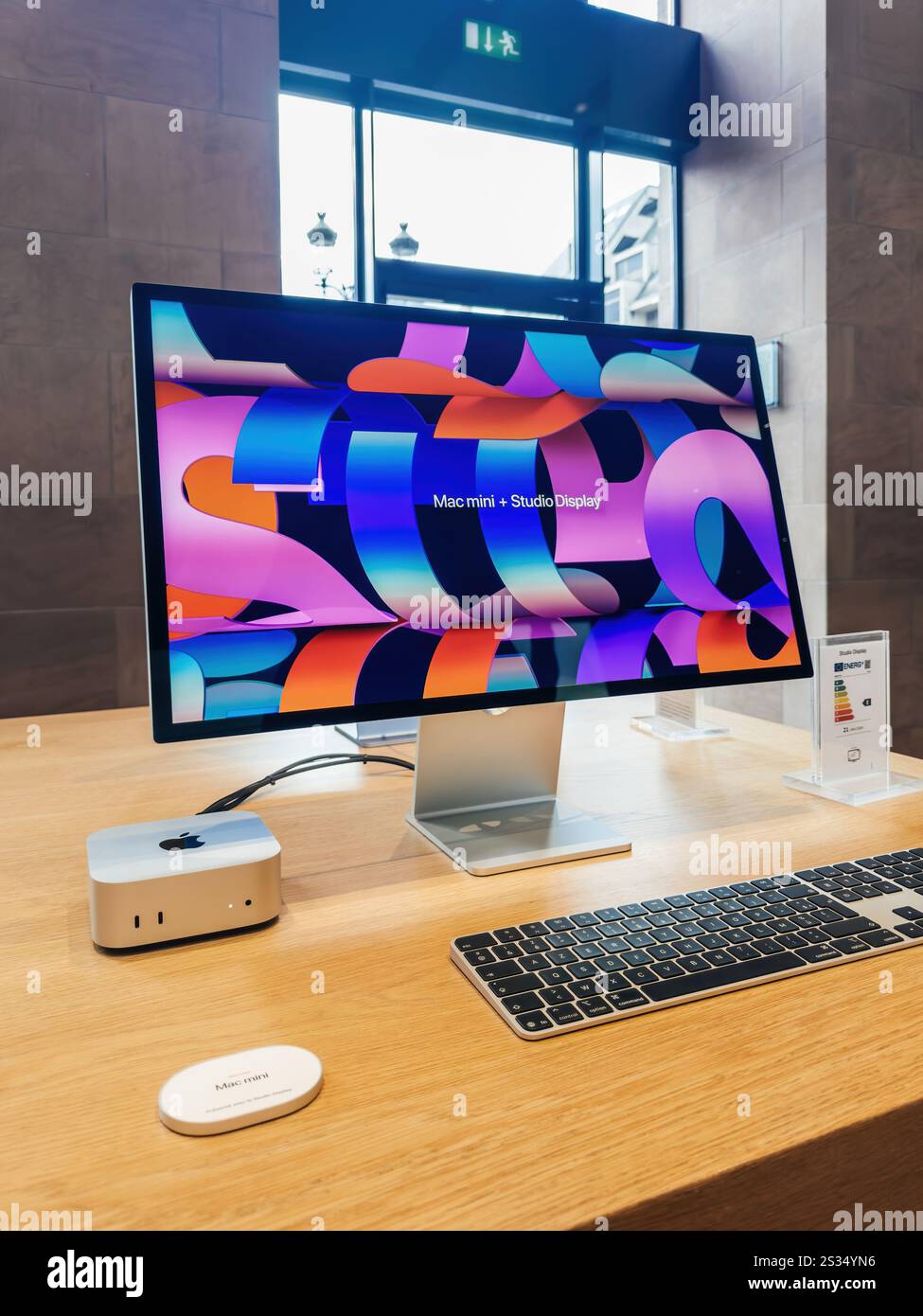 Paris, Frankreich - 10. Dezember 2024: Ein schlankes Mac Mini-Setup mit Studio-Display mit brillanter Grafik, Tastatur und Werbeletiketten, präsentiert auf einem Holztisch in einem modernen Verkaufsraum. Stockfoto