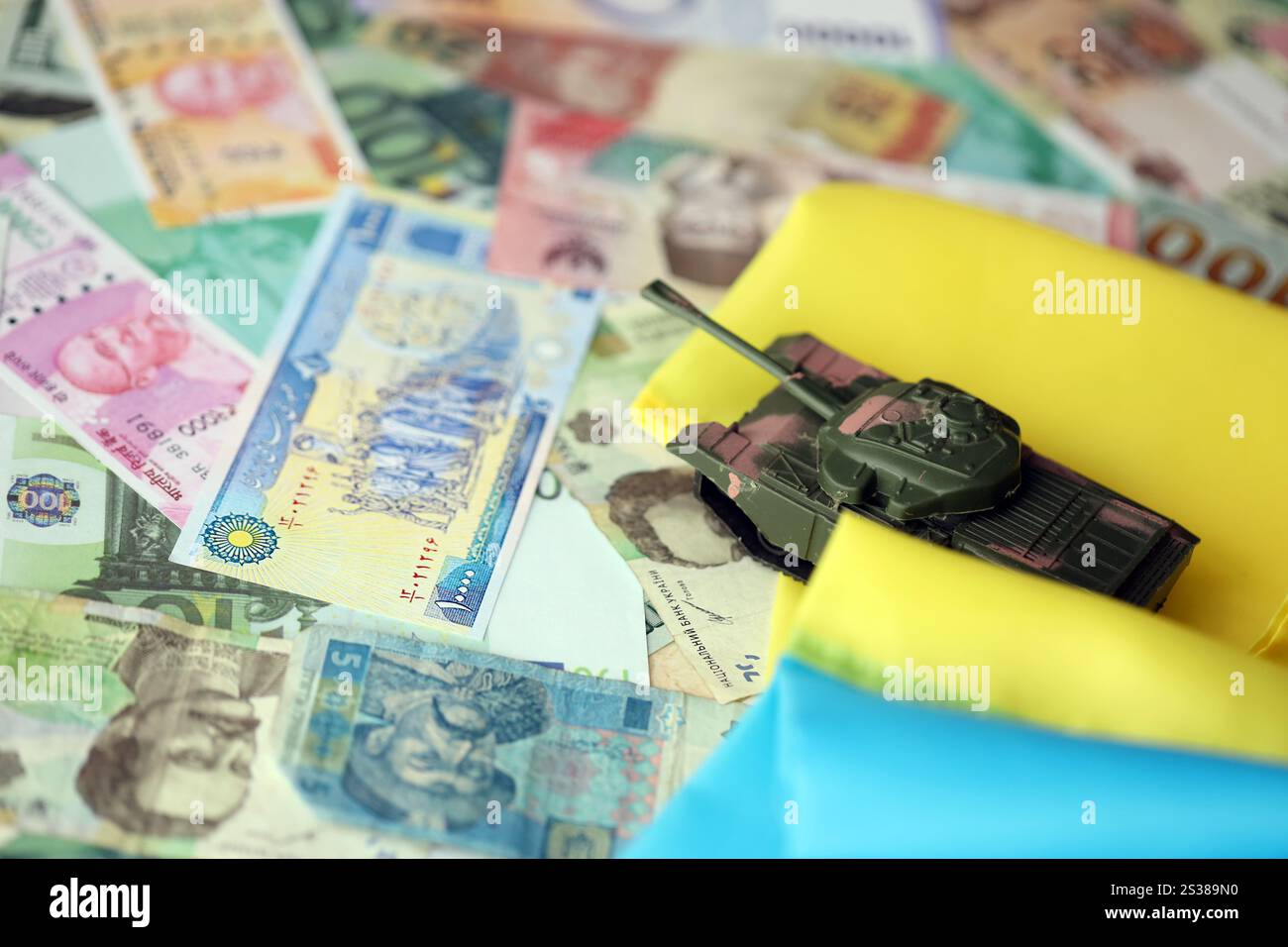 Spielzeugtank auf ukrainischer Flagge auf vielen Banknoten unterschiedlicher Währung. Hintergrund der Kriegsfinanzierung und des Preises für militärische Unterstützung in der Ukraine. Spielzeugtank an Stockfoto