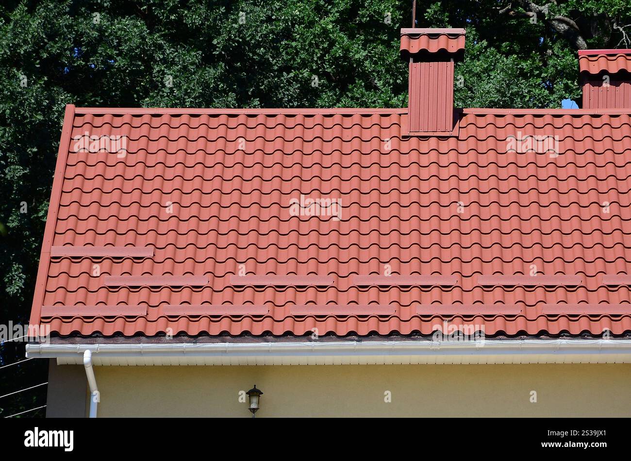 Hochwertiges, rotes Metallziegeldach eines Hauses vor grünem Baumhintergrund. Hochwertiges Dach aus rotem Metallfliesen eines Hauses Stockfoto