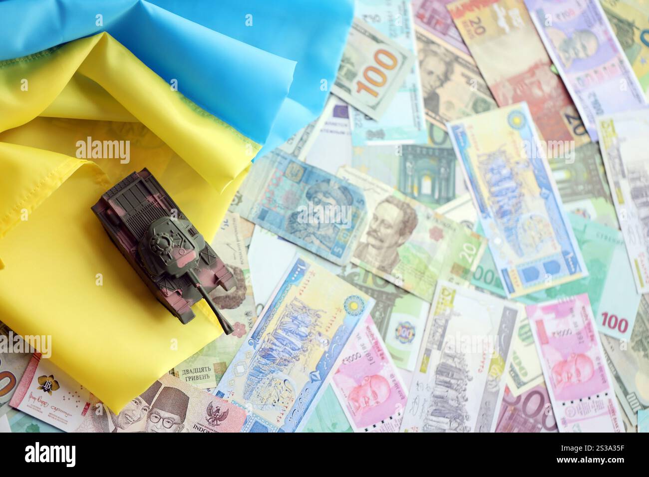 Spielzeugtank auf ukrainischer Flagge auf vielen Banknoten unterschiedlicher Währung. Hintergrund der Kriegsfinanzierung und des Preises für militärische Unterstützung in der Ukraine. Spielzeugtank an Stockfoto