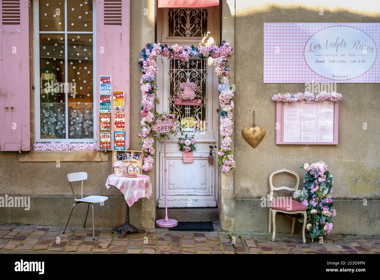 Die charmante rosafarbene Fassade und der mit Blumen dekorierte Eingang des Teehauses Les Volets Roses salon de thé in Bayeux, Normandie, Frankreich Stockfoto