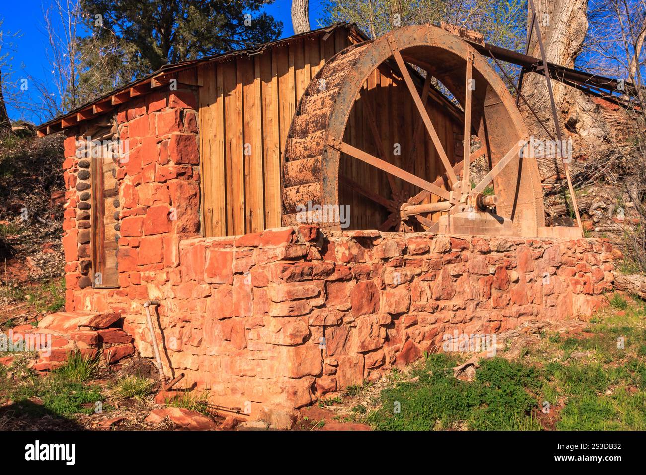 Ein kleines Gebäude mit einem Holzrad an der Seite. Das Rad ist alt und verrostet. Das Gebäude ist aus Stein und hat ein rustikales Aussehen. Das Gras ist Stockfoto