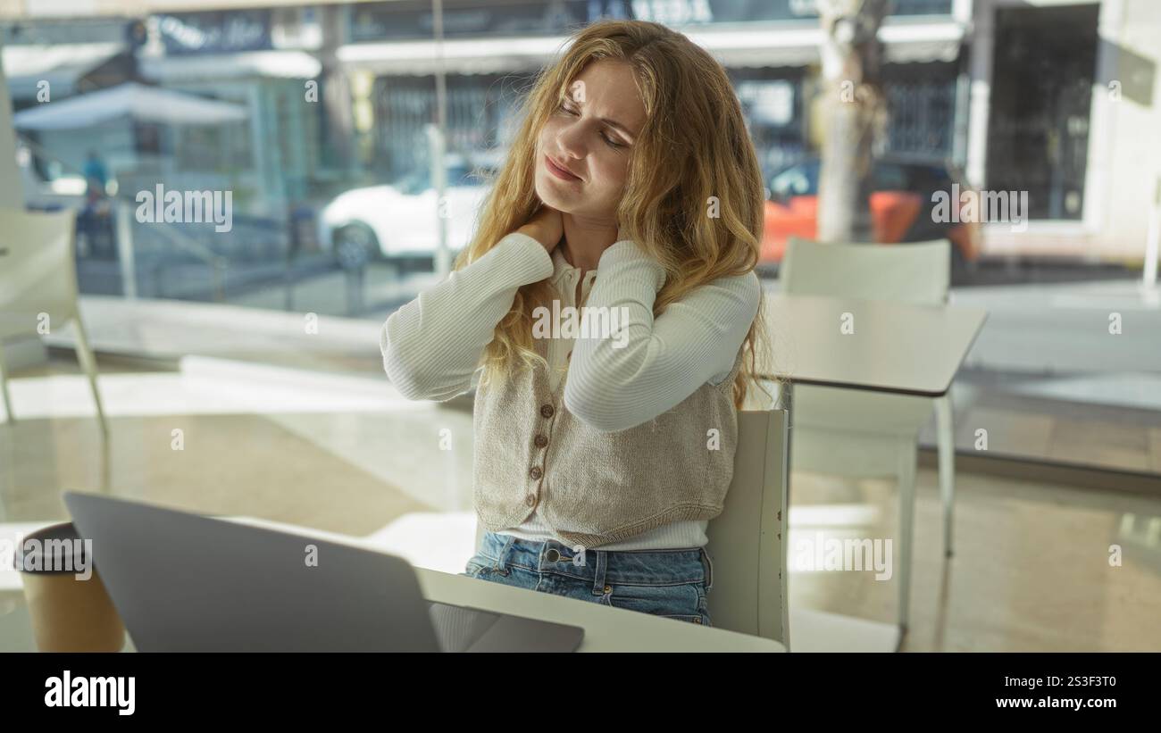 Frau im Büro mit Laptop, die Nackenbeschwerden hat, hebt den Stress am Arbeitsplatz hervor und bietet eine Inneneinrichtung mit sanftem Licht und Kaffee Stockfoto